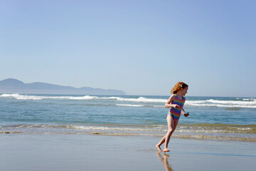 Fototapeta premium Happy little redhead girl running along the ocean shore.
