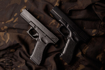 Two black pistols are lying parallel on a camouflage soldier's jacket. A firearm that makes you think about self-defense. Weapons Day. Against violence. Top view