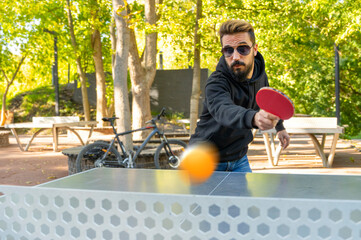 Playing table tennis outdoors at the open air sport ground. Stylish man in sunglasses with beard...