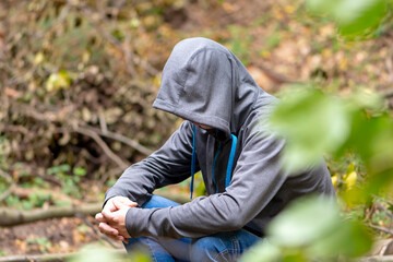 A man is sitting in a jacket with a hood hiding his face in the forest under a tree. Perhaps he is a lost tourist, a fugitive, an emigrant, a border trespasser, a fugitive from justice.