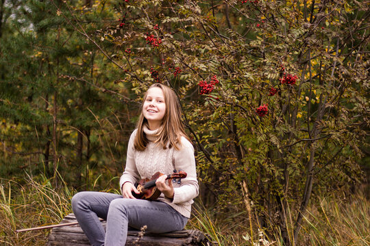 A beautiful teenage girl with long blonde hair sits on a log in the park and holds a violin in her hands, looks happ