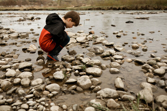 Young boy crouching in a river bed surrounded by rocks in a red jacket
