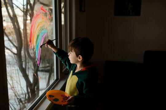 Young boy in costume painting a rainbow on a window in a living room