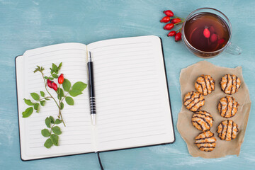 Coconut macaroons on a textured blue background, empty booklet with a pen, cup of tea with red hip rose