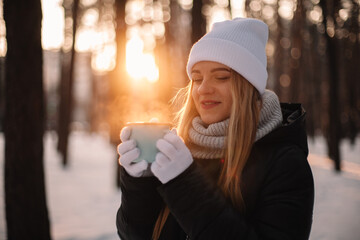 Young woman holding cup of tea standing at park in winter at sunset