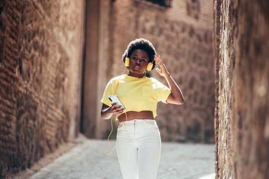 Portrait of black girl with afro hair listening to music with headphones and her mobile phone while walking down an old city street.