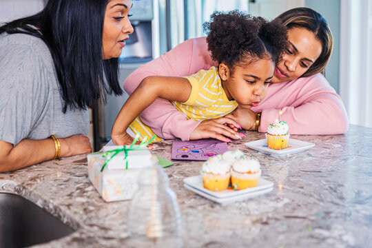 Girl blowing candle on cupcake while celebrating birthday with mother and grandmother at home
