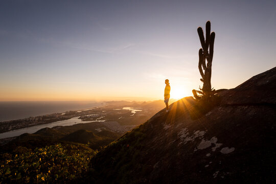Man standing on rocky mountain edge next to big cactus on sunset