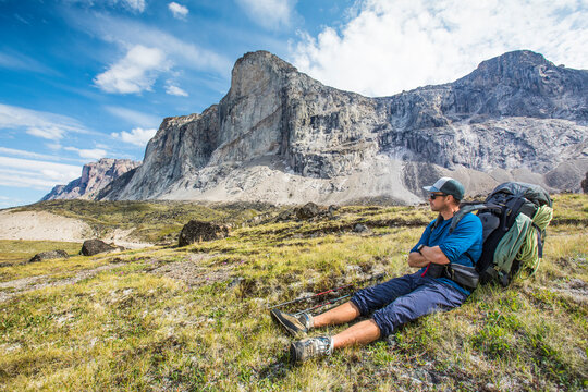 Backpacker resting, sitting in grassy meadow below mountain summit.
