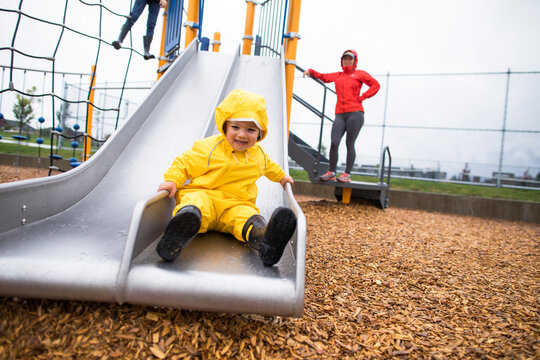 Happy boy sliding on slide at local park, mother watching.