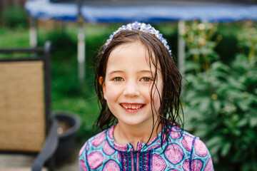 A smiling child stands with direct gaze, wet hair, and a purple crown