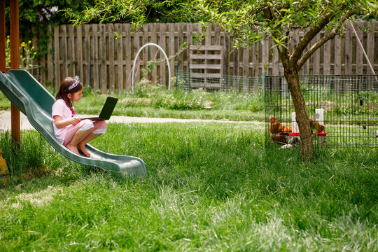 A small girl sits on playset in garden with computer in virtual school