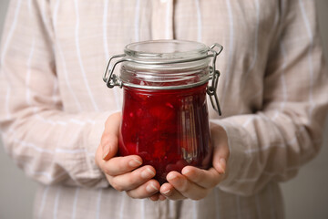 Woman holding glass jar with sweet cherry jam on grey background, closeup
