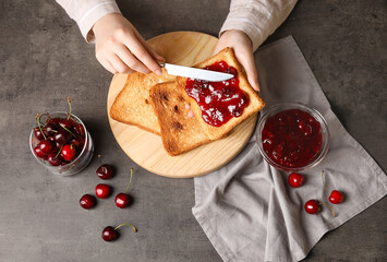 Woman spreading sweet cherry jam onto toast on dark background