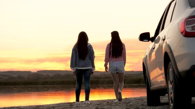 Girls drivers stopped at campsite. Free women travelers stand next to car on beach enjoying sunset in park hug and rejoicing. Girlfriends are enjoying trip in car. Travel on vacation by car, adventure