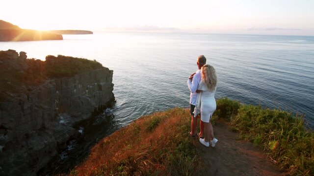 A blonde pregnant woman in white dress is hugging her husband gently with love. They both are stading on the cliff edge above the sea and enjoying beautiful dawning