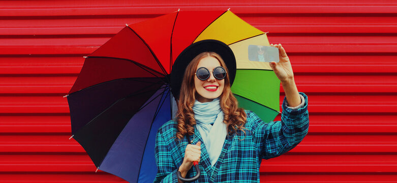 Portrait of happy smiling young woman taking a selfie picture by phone holding colorful umbrella on red background