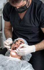 Mujer con cofia descartable, abre su boca para ser atendida por un odontólogo con un espejo y una pinza en el consultorio - obrazy, fototapety, plakaty