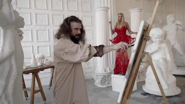 a bearded man with long hair and a shirt on naked body paints a picture on an easel in a white space. a blonde in a red dress froze in the background out of focus