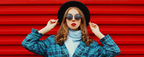 Portrait of beautiful young woman wearing a black round hat, blue knitted coat on red background