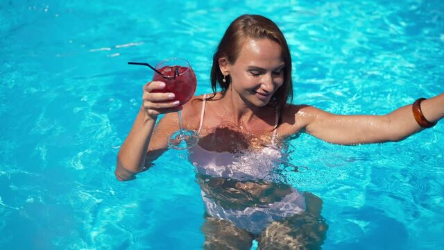 Tanned beautiful slim woman in pink bikini dancing in swimming pool in slow motion smiling. High angle view of gorgeous confident Caucasian tourist with cocktail glass enjoying leisure in sunshine