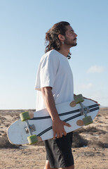 Young adult skater profile portrait standing outdoors holding surfskate. He has long black hair and he is wearing a white shirt. © 1 MEDIA