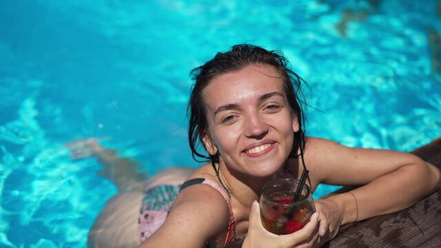 High angle view of smiling satisfied young woman drinking cocktail smiling in slow motion at poolside in blue swimming pool water. Portrait of relaxed carefree joyful tourist posing outdoors