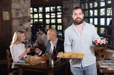 Polite bearded waiter offering delicious dishes for tasting in cosy rustic restaurant ..