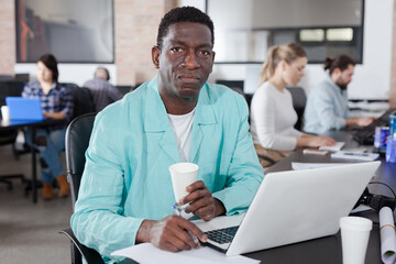 Portrait of smiling successful African businessman sitting with laptop in modern coworking space