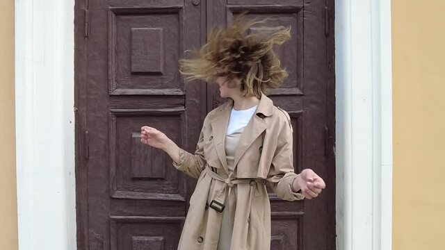Young smiling millennial woman dressed in an autumn coat dancing and shaking hair near the door of an old building.