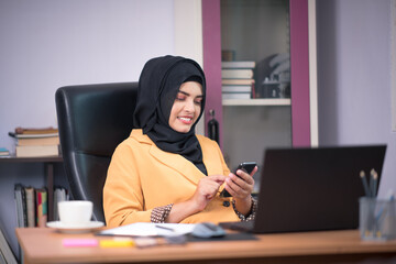 Fototapeta na wymiar An office worker for an Asian Muslim woman sitting in front of a laptop computer at her desk and talking on a mobile phone and working at the office.