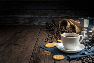 Hot coffee in a white coffee cup and many coffee beans placed around and sugar on a wooden table in a warm, light atmosphere, on dark background.