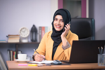 An office worker for an Asian Muslim woman sitting in front of a laptop computer at her desk and talking on a mobile phone and working at the office.
