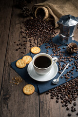Hot coffee in a white coffee cup and many coffee beans placed around and sugar on a wooden table in a warm, light atmosphere, on dark background.