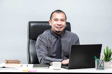 Asian male office worker sitting in front of a laptop computer at a desk and smiling at the office