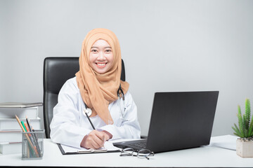 The Asian Muslim woman doctor was sitting at the patient's table and looked at the camera with a smiling and welcoming face.