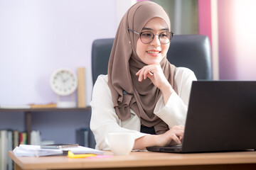 Asian Muslim woman office worker sitting in front of a laptop computer at a desk and smiling happily at the office.