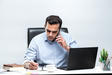 Asian male office worker sitting in front of a laptop computer at a desk and talking on a mobile phone and taking notes at the office.
