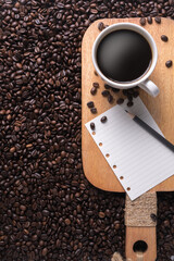 Coffee in a white cup with notes and pencils placed on a wooden tray that is surrounded by many coffee beans, with copy space.