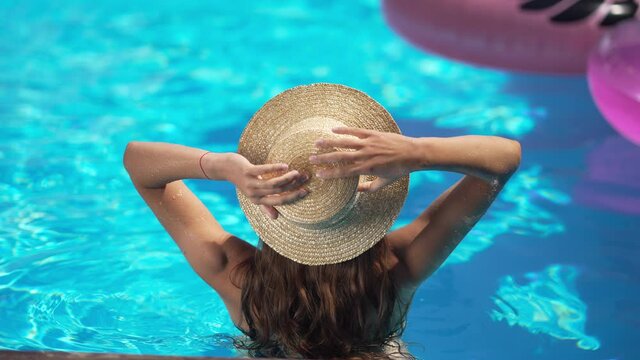 Back view of slim Caucasian young woman putting on straw hat in slow motion standing in azure blue water in swimming pool outdoors. Relaxed happy satisfied tourist in sunshine at resort