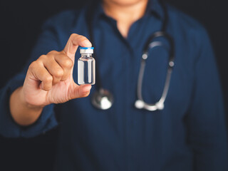A doctor is holding a vaccine bottle while standing in the studio with a black background. Vaccine...