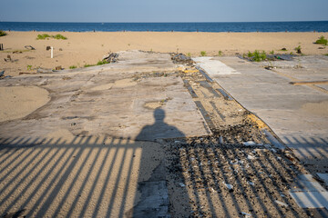 Shadows of person and fence overlooking entrance to beach from boardwalk. Sand & ocean in distance. 