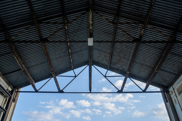 View looking up to industrial building roof from inside, sky & clouds behind.