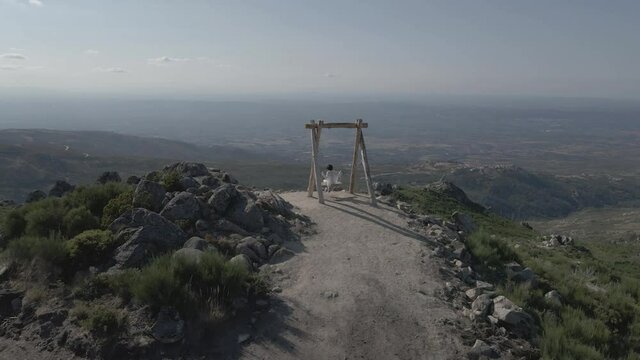 Drone pulls back from a beautiful woman swinging on a dirt path into an establishing shot overlooking a valley.