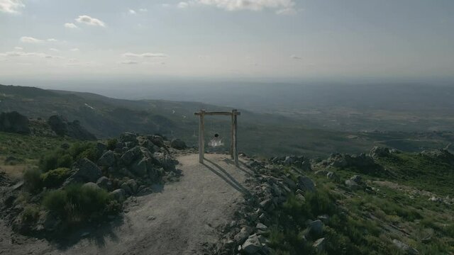 Centered on a swing with a woman on a white jacket swinging in the wind as the drone pulls back into an overview.