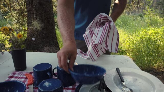 Low Angle Static Shot Of Man Drying Camping Dishes Outdoors At Campsite.