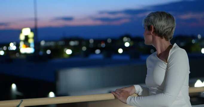 Beautiful slim slender sexy short blonde haired caucasian women in white dress looking over rail towards pink and blue fading sunset and city lights. In Cinema 4K (4096x2160) 30fps slowed from 60fps.