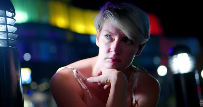 Pretty beautiful girl lady women with short pixie cut blonde hair sitting in front of gay lesbian pride colored rainbow lights on building at night. In Cinema 4K (4096x2160) 30fps slowed from 60fps.