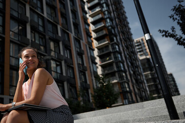 Portrait of a gorgeous African American woman talking on mobile phone, looking away, posing to camera on urban high buildings background. Business, office, freelance, remote work, online concept