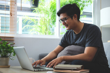Smiling Asian man surfing internet with computer laptop in living room.